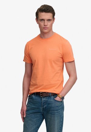 Joven con camiseta naranja, vaqueros azules, cinturón marrón y reloj de pulsera, de pie con una mano en el bolsillo frente a un fondo blanco.