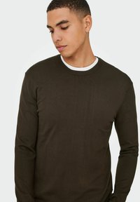 Ciemnozielony sweter z długim rękawem, okrągłym dekoltem i subtelną teksturą ściągacza, nałożony na biały T-shirt z okrągłym dekoltem. 