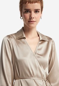 Beige satinomslagen klänning med krage, långa puffärmar och knytning i midjan. Slät textur med en lätt lyster, med en djup utrigning.