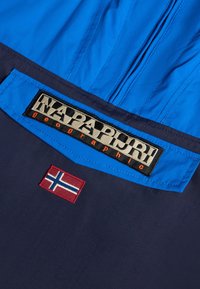 Napapijri-jas in een blauw en marineblauw kleurenpalet, met een opvallende logo-uptak en een geborduurd Noorwegen-vlag detail. Soepele textuur.