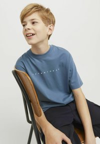 Sinine puuvillane T-särk lühikeste varrukatega ja ümmarguse kaelusega, mille ees on keskelt valge tekst "JACK&JONES". Mudel istub toolil.