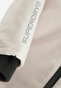 Manche de veste beige clair avec un poignet côtelé noir, arborant un logo "SUPERDRY®" gris bien en vue, et une fermeture éclair noire sur le côté.