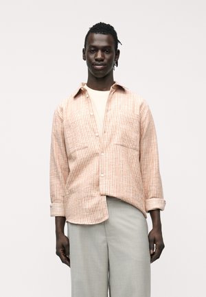 Άνδρας με σκούρο δέρμα που φοράει ανοιχτό μπεζ πουκάμισο κάτω από πουκάμισο με ροζ ρίγες και ανοιχτό γκρι παντελόνι, στέκεται μπροστά σε απλό λευκό φόντο.