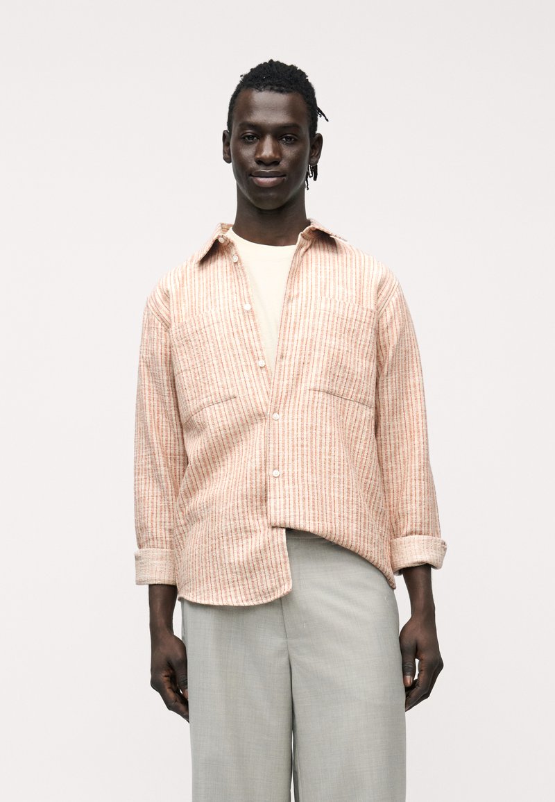 Άνδρας με σκούρο δέρμα που φοράει ανοιχτό μπεζ πουκάμισο κάτω από πουκάμισο με ροζ ρίγες και ανοιχτό γκρι παντελόνι, στέκεται μπροστά σε απλό λευκό φόντο.