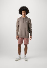 Lichtbruine hoodie met een voorkantzak en trekkoord, gecombineerd met roze shorts en witte sneakers, tegen een effen achtergrond.