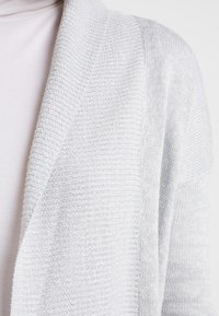Jasnoszary kardigan o teksturowanym wzorze dzianiny, z otwartym przodem i lekko luźnym krojem, wykonany z miękkiej i lekkiej tkaniny.
