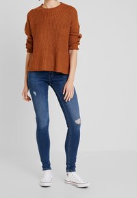 Maglione lavorato a maglia arancione rustico con trama a costine, abbinato a jeans skinny blu con dettagli distrutti e sneakers di tela bianche.