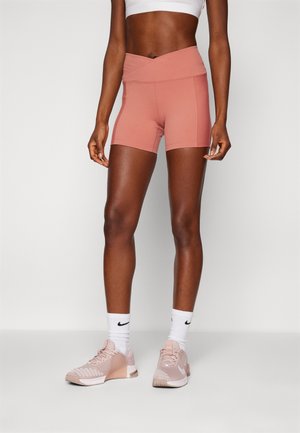 Personne en forme portant un short cycliste taille haute mauve, un soutien-gorge de sport blanc, des chaussettes Nike blanches et des baskets Nike mauves, debout sur un sol uni.