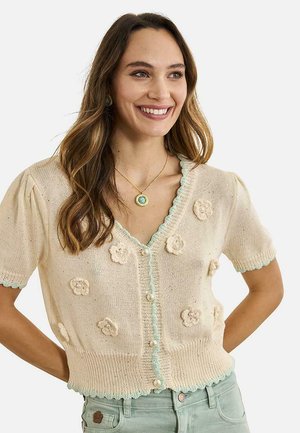 Mujer sonriente con cabello castaño ondulado que lleva un cárdigan de punto crema de manga corta con apliques de flores y botones de perla, combinado con pantalones verde claro.