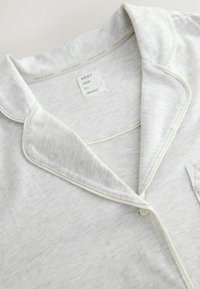 Hellgraues Pyjama-Oberteil aus weichem Stoff, mit klassischem Kragen, weißen Paspeln und einer Brusttasche mit einem Etikett, auf dem "NEXT" steht.