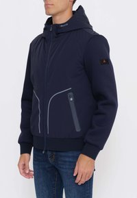 Uomo che indossa una giacca con cappuccio blu navy con cerniera frontale e tasca sul petto, abbinata a jeans di denim blu, su uno sfondo semplice.