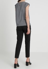 Femme portant un haut sans manches gris à motifs, un pantalon noir slim-fit et des chaussures ouvertes à talons blocs noires, se tenant de dos.