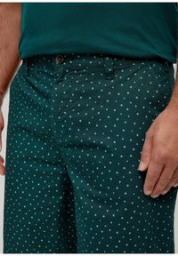 Donkergroene katoenen broek met een subtiel patroon van kleine lichtblauwe vormen. Voorzakken en een knoopsluiting zijn zichtbaar.