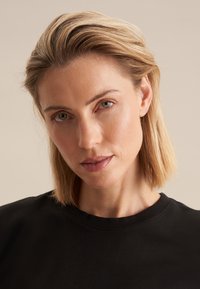 Zwart T-shirt met een ronde hals, gedragen door een persoon met halflang blond haar. De stof lijkt glad te zijn met een minimalistisch ontwerp.