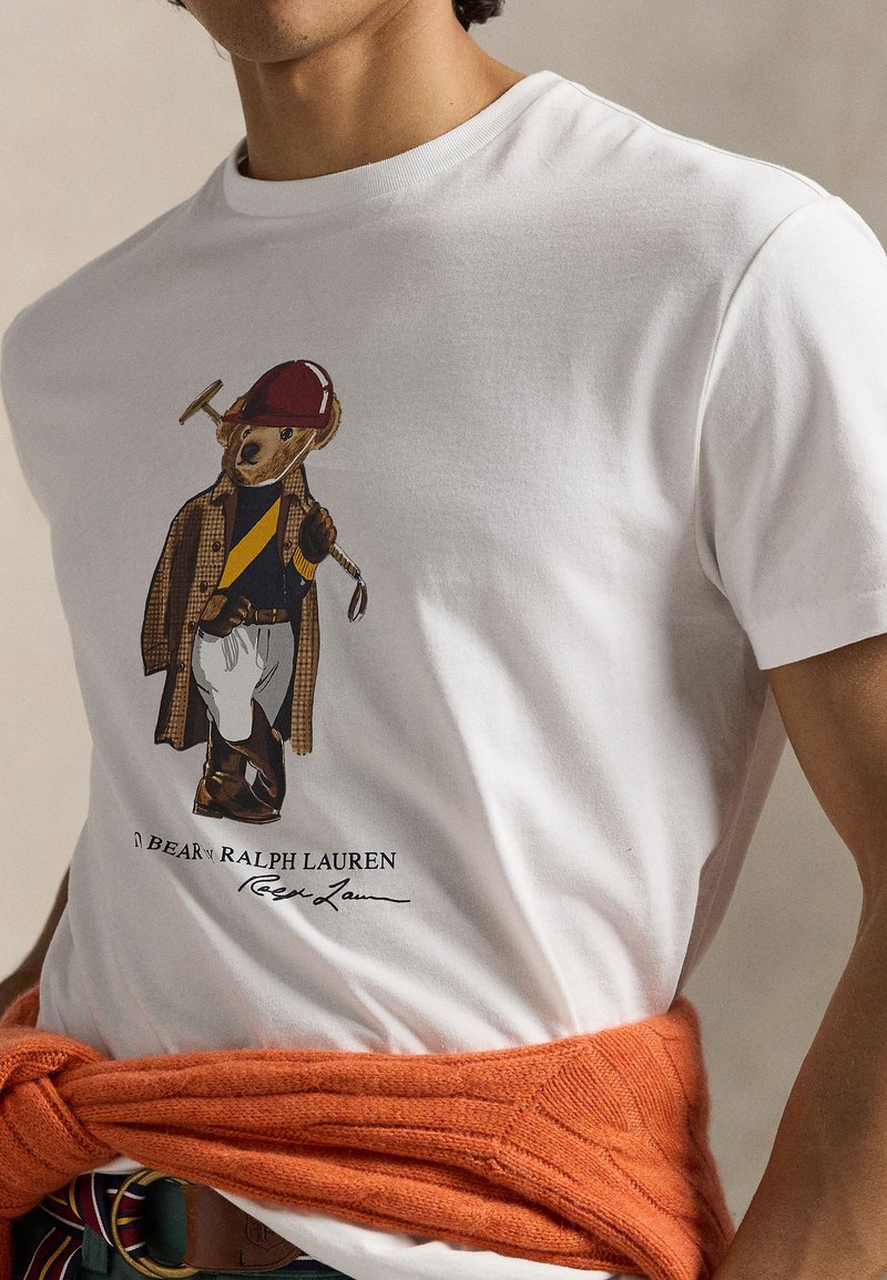 Weißes Baumwoll-T-Shirt mit einem Bären in einem karierten Mantel und rotem Schutzhelm, der einen goldenen Schläger in der Hand hält. Orangefarbener Strickpullover, in der Taille gebunden.