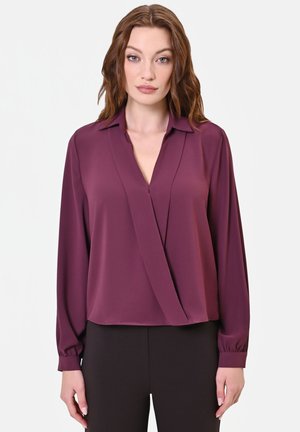 Donna dalla pelle chiara e capelli castani mossi, che indossa una camicetta viola scuro a maniche lunghe e pantaloni neri, in piedi davanti a uno sfondo bianco semplice.