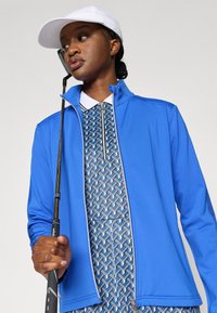 Zils rāvējslēdzēja džemperis ar augstu apkakli, valkāts virs rakstainas polo kleitas ar ģeometriskām formām zilā un baltā krāsā. Rokā tur golfa nūju.