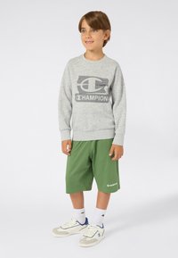 Felpa grigia con logo "CHAMPION" in un design a blocchi, abbinata a pantaloni corti verdi e sneaker bianche con dettagli blu.