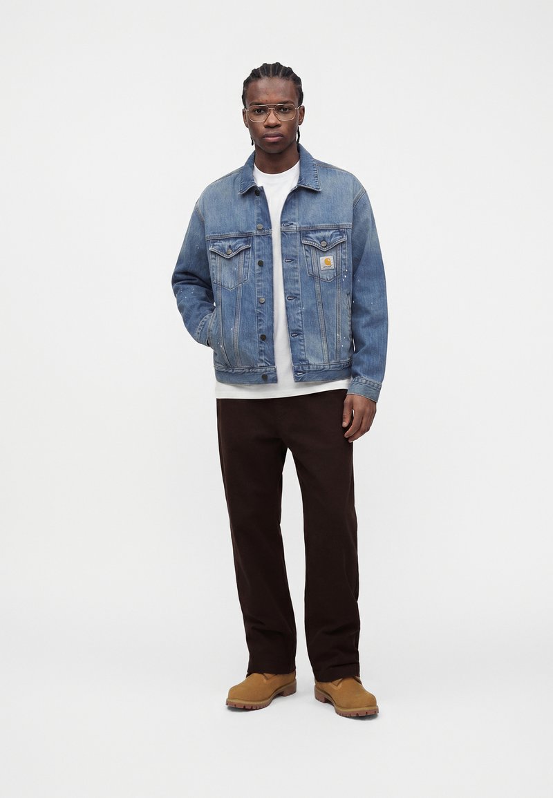 Uomo con capelli intrecciati che indossa occhiali trasparenti, giacca di jeans blu, camicia bianca, pantaloni scuri e stivali marroni, in piedi su sfondo bianco.