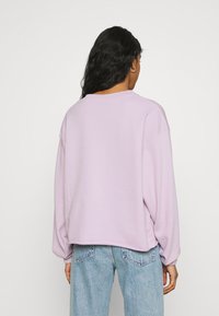 Kvinna med långt mörkt hår bär en stor ljuslila sweatshirt och ljusblå jeans, sedd bakifrån mot en enkel bakgrund.