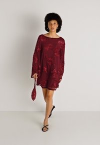 Βουργουνδικό φόρεμα με λουλουδάτη κεντητική, μακριά μανίκια και άνετη εφαρμογή, συνδυασμένο με τσάντα σε ματς χρώμα και μαύρες σανδάλες.