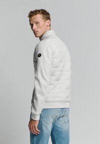 Witte gewatteerde jas met een hoge kraag, voorzien van zijzakken, contrasterende gestructureerde mouwen en een hexagonaal logo op de linkerarm.