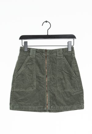 Groene corduroy minirok met ritssluiting aan de voorkant, riemlussen en twee grote voorzakken, hangend aan een zwarte hanger.