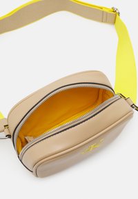 Borsa a tracolla in pelle beige con una tracolla gialla, dotata di chiusura zip e un luminoso interno giallo. Logo impresso sul fronte.