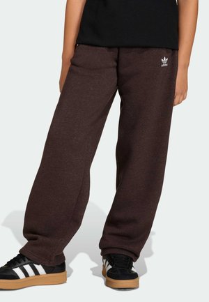 Pantalones deportivos marrones de tela suave, con un corte holgado y el logo blanco de Adidas en el frente. Combinados con zapatos negros con rayas blancas.