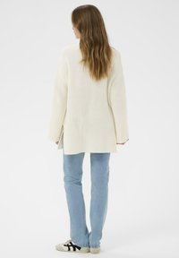 Cardigan blanc à côtes avec une coupe décontractée, porté sur un jean bleu clair et des baskets beiges avec des accents noirs. Le design présente des fentes latérales.