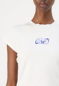 Fehér rövid ujjú pamut póló, a mellkasán kék, írott stílusú "OBEY" logóval és nyers szélű nyakkivágással.