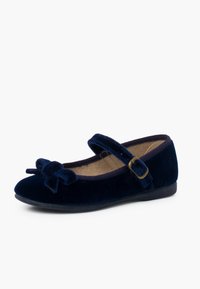 Zapato plano de terciopelo azul marino con punta redondeada, que presenta un detalle de lazo y una correa con una hebilla dorada. Superficie suave y texturizada y tacón bajo.