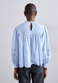 Vaaleansininen pusero, jossa on avonainen selkä, pleatattu muotoilu ja pussitettu hiha. Sisältää herkkiä kirjontateoksia ja rento, väljä istuvuus.