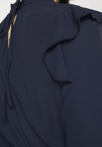 Marinblus med volangdetaljer på axlarna och knytning i ryggen. Tillverkad av lättviktsmaterial med en slät yta.