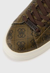 Καφέ πάνινο αθλητικό παπούτσι με διακοσμητικά σχέδια, σκούρα κορδόνια και λογότυπο «GUESS». Λαστιχένια σόλα με στρογγυλεμένο σχέδιο μύτης.