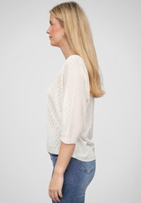 Lys beige strikkegenser med perforert design, vide trekvartermiddermer og avslappet passform, kombinert med blå jeans.