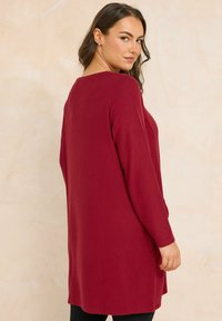 Vrouw met lang donker haar, gekleed in een lange mouw, knielang bordeauxrood trui, die over haar schouder kijkt tegen een beige achtergrond.