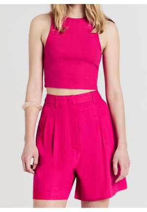 Femme portant un crop top sans manches rose vif et un short rose taille haute ajusté, debout devant un fond blanc uni.