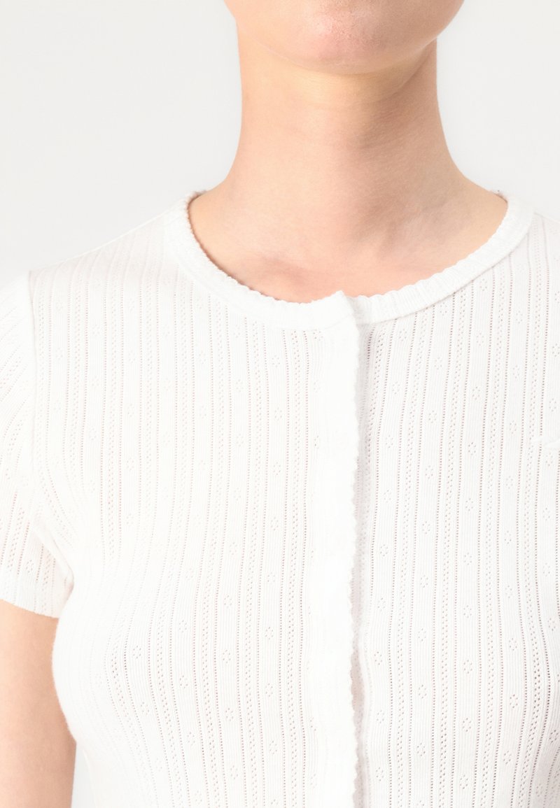 Biała, prążkowana bluzka dzianinowa z krótkimi rękawami, okrągłym dekoltem w falbany, teksturowanym wzorem z małymi wycięciami oraz zapięciem na guziki z przodu.
