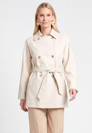 Kvinde med langt blondt hår iført en beige trenchcoat med bælte og store guldringe, stående foran en ensfarvet lys baggrund.