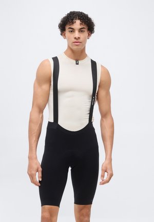 Fiatal férfi göndör hajjal, bézs ujjatlan felsőt és fekete kerékpáros nadrágot, lólábbal viselve, világos egyszínű háttér előtt áll.