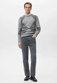 Suéter gris con un patrón argyle y acentos blancos, combinado con unos jeans rectos de color gris oscuro y zapatos negros pulidos. El modelo se encuentra en un fondo simple.