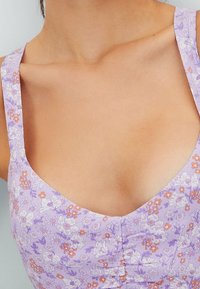 Gros plan sur une femme portant une robe sans manches lilas à fleurs avec un détail froncé au niveau de la poitrine.