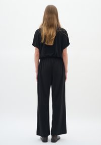 Svart bredbenad jumpsuit med elastisk midja, korta dolmans ärmar och en mjuk tygtextur. Stylad med platta svarta skor.
