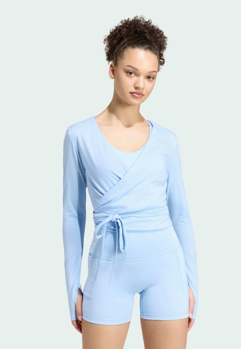Jeune femme aux cheveux bouclés porte un haut bleu clair à manches longues style cache-cœur et un short ajusté assorti sur fond uni.