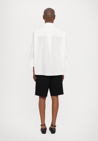 Hvit oversized skjorte med bakfold, lange ermer og rund hem, kombinert med svarte shorts og svarte spiked sko.