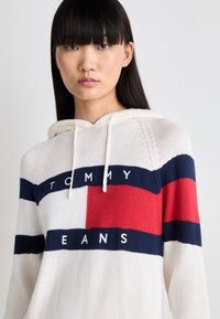 Pullover a maglia con cappuccio di colore crema con strisce blu navy, rosse e bianche. Presenta la scritta "TOMMY" e "JEANS" stampata in blu su una toppa bianca.