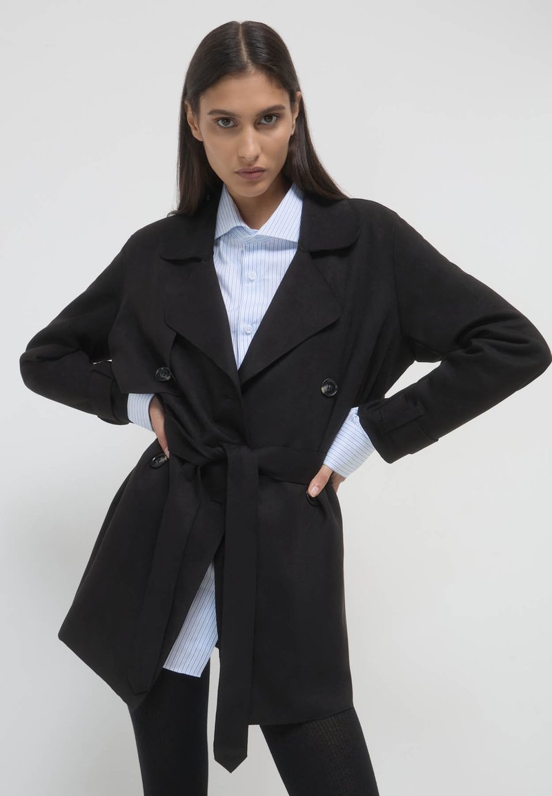 Cappotto trench nero con cintura in vita, ampi rever e due bottoni, indossato sopra una camicia a righe blu chiaro. Tessuto liscio.