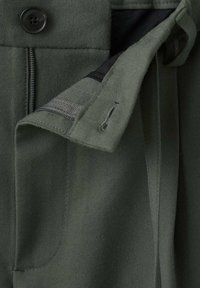 Tela de color verde oliva con una textura suave, que presenta un cierre de cremallera y botón. Incluye un bolsillo y un detalle de lazo en la cintura.