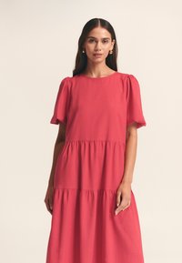 Robe rouge à étages avec un col rond et des manches courtes bouffantes. Confectionnée dans un tissu texturé, avec une taille froncée.